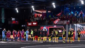 Jugadores de dos equipos frente a frente en una pista de fútbol indoor de la Kings League, con público animando y banderas en la grada.
