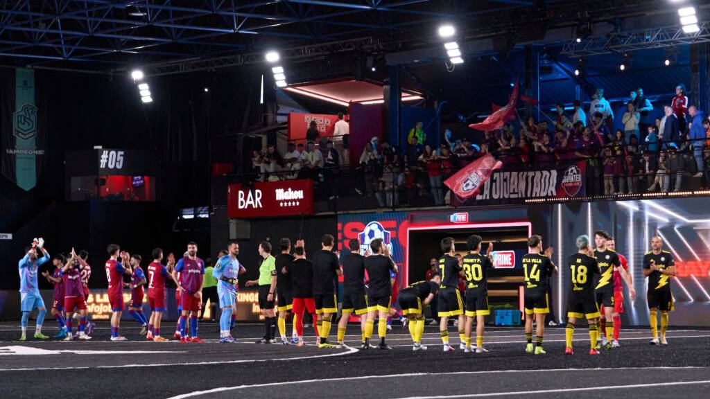 Jugadores de dos equipos frente a frente en una pista de fútbol indoor de la Kings League, con público animando y banderas en la grada.